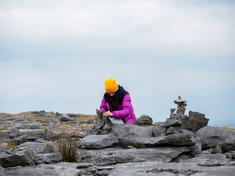 Woman in purple jacket and yellow beanie arranges stones on rocky terrain. She focuses intently on balancing stones near a stone cairn. Overcast sky adds calm atmosphere to open, rugged landscape