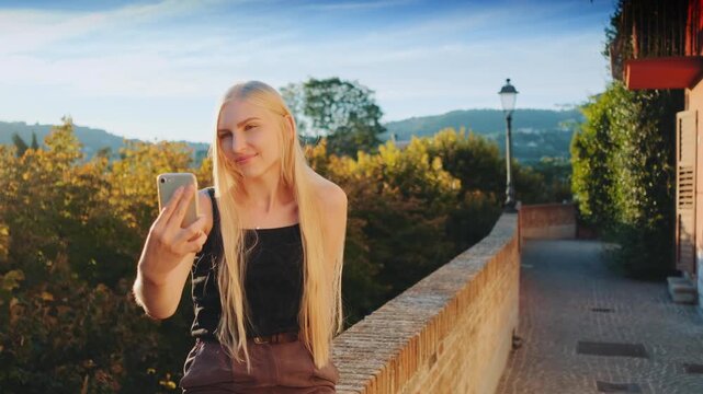 A blonde woman in a black tank top and brown shorts takes a selfie while sitting on a stone wall overlooking a lush forest and rolling hills during daytime.