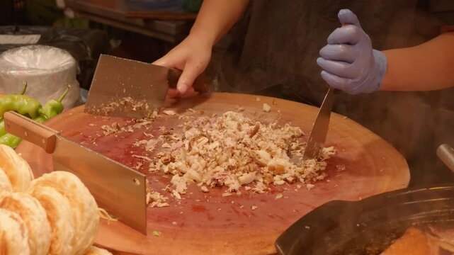 Cook minces cooked meat on a large wooden cutting board, preparing filling for flatbread at a food stall.