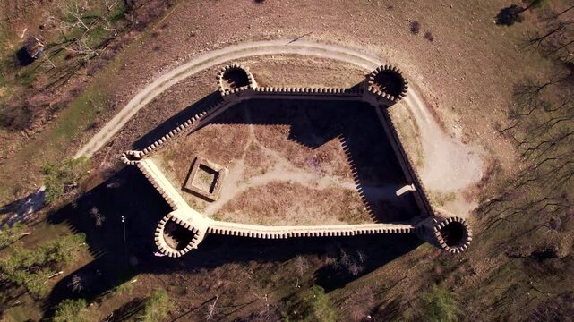 Stone fortress forms a near rectangular outline in a top down drone view with four round towers an empty courtyard and small inner foundation while a pale path bends around its outer walls