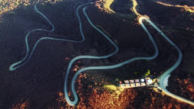 Mountain road makes repeated switchbacks through leafless trees with bright curves against the dark slope while small roadside buildings line the lower junction in a high overhead view