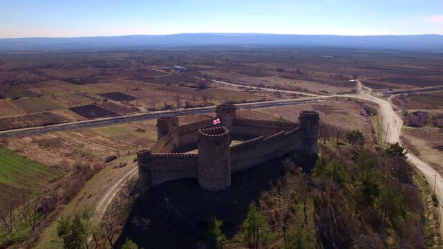 Stone fortress grows larger during an aerial approach over brown fields revealing its four towers open inner court and Georgian flag with a paved road and pale dirt junction beyond the walls