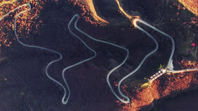 Winding mountain road forms a long series of linked hairpins across a dark leafless slope while patches of afternoon sun mark the ridges and a small roadside cluster remains at the far edge