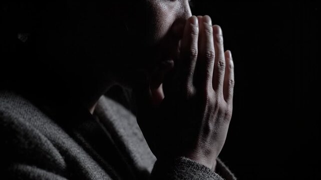 Person bows head with clasped hands in prayer against a dark background. Spiritual reflection and devotion. Faith, religion and worship. Contemplative close-up video.