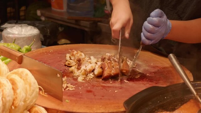 A chef wearing gloves cuts cooked meat with a cleaver on a wooden chopping board in a food stall.