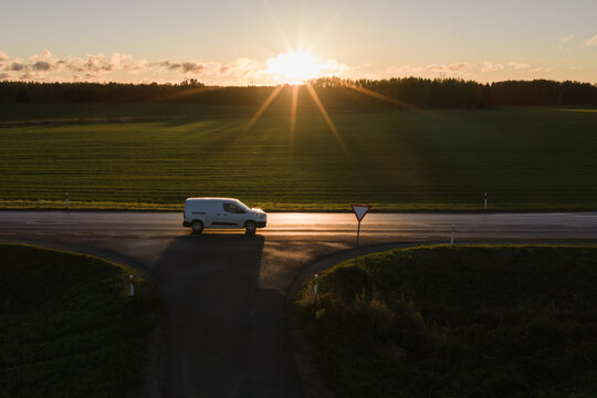 Aerial drone view of white van waiting at intersection with yield sign on rural country road against bright sunrise horizon in morning