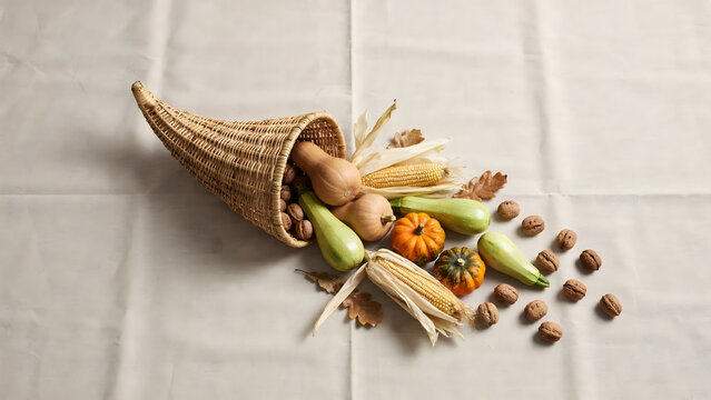 A woven cornucopia basket filled with various autumn vegetables and nuts resting on a plain white fabric background, for harvest display