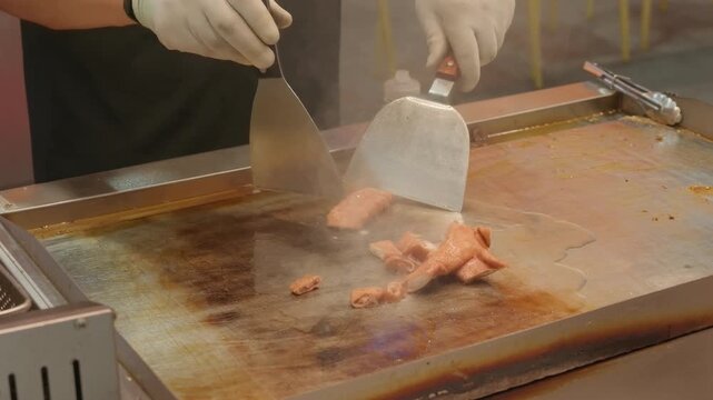 A chef wearing gloves prepares meat on a commercial stainless steel griddle using a knife and spatula in a kitchen setting.