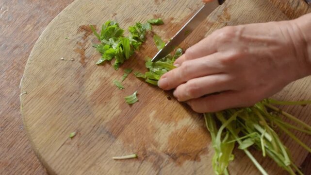 Close up overhead view of woman hands chopping fresh salad greens on round wooden cutting board in rustic kitchen with natural light, showing healthy food prep and simple home cooking