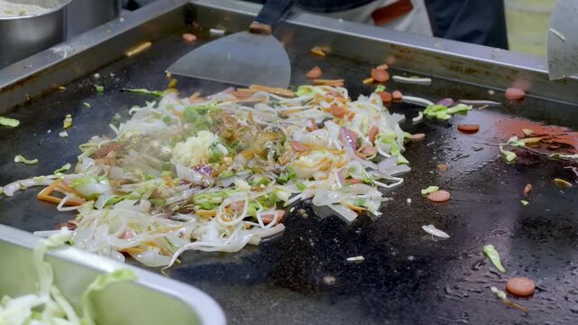 Noodles and mixed vegetables being stir-fried on a hot flat griddle, with cooking utensils and a chef's gloved hand visible.