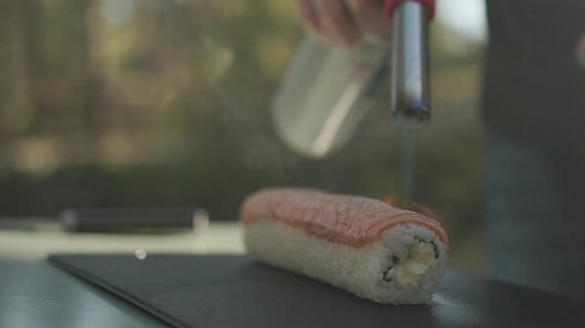 Close cooking process as a sushi chef torches salmon covering a prepared roll using intense flame to brown the fish surface and enhance the fresh seafood topping