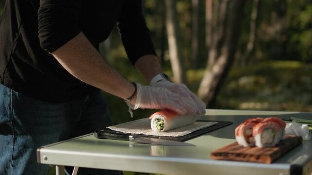 Professional sushi maker in protective gloves lays thin pieces of fresh fish neatly over a roll showing the detailed process of preparing traditional Japanese sushi