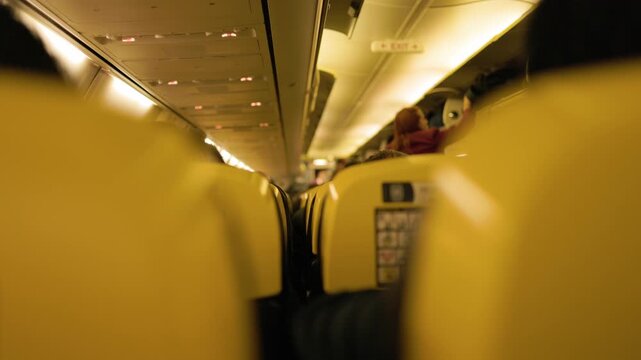 View through a passenger airplane cabin during flight showing people seated in rows beneath warm lights capturing the everyday experience of commercial air transportation