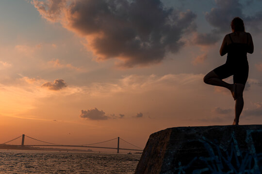 Woman balancing on one leg in tree pose at sunset. A woman balances on one leg facing the ocean in New York with a suspension bridge behind her.