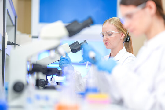 Blonde female scientist pipetting at a microscope while a brunette colleague works in the foreground during a microbial assay for an AGI-guided pathogen detection study