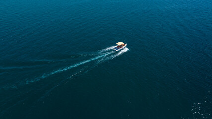 Aerial top-down view of a small motorboat cruising through deep blue waters, leaving a white foamy wake behind as it glides across the serene open surface. © Stefano Tammaro