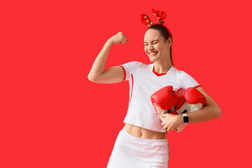 Holiday fitness and strength, happy woman with boxing gloves and reindeer antlers celebrating Christmas health goals on a solid red background © Pixel-Shot