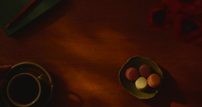 Moody overhead flat lay of a cup of black coffee and colorful macarons on a wooden table, with hands gently holding the saucer and warm evening light creating an intimate caf&eacute; atmosphere.