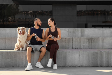 Fototapeta premium Healthy lifestyle and fitness, young couple with dog resting after workout and drinking water on concrete steps in urban environment