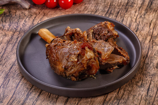 Roasted lamb shank with exposed bone and tender meat on dark ceramic plate with cherry tomatoes background rustic dinner