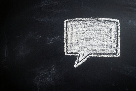 White chalk speech bubble on black blackboard background. Empty talk balloon symbol for communication message social media text. Hand drawn cloud shape provides copy space for ideas, discussion,