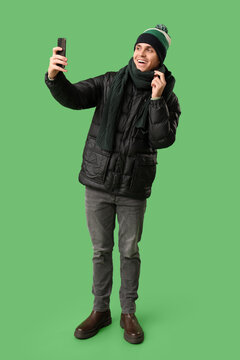 Social media influence and digital connection, young man taking a selfie with a smartphone wearing winter clothes on a green background