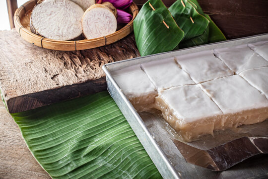 Traditional Thai tako taro pudding dessert square cut topped with thick coconut milk cream in aluminum tray next to banana leaf parcels and fresh sliced taro root. Asian sweet food snack.