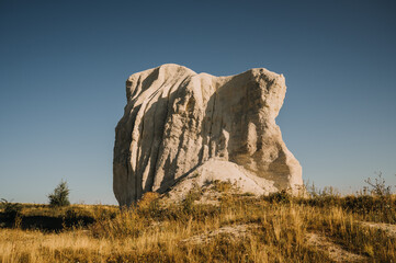 white limestone rock spire rises from dry grassland, rugged texture and shadows under clear blue sky in sun © Vulp
