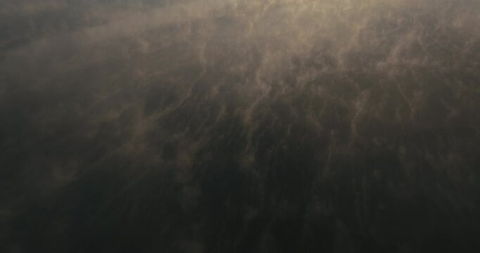Drone flies above a misty lake as drifting fog creates mesmerizing abstract swirling patterns on the water, then tilts upward to reveal a pine forest and clear cut area.