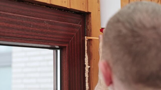 Worker applies construction foam around wooden window frame during insulation repair