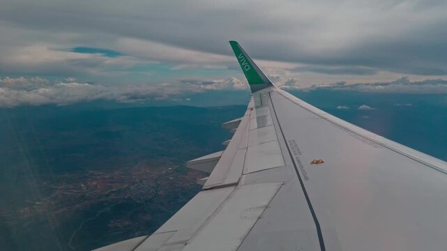 Airplane wing and green winglet seen from passenger window during flight above clouds and distant landscape. Aircraft wing detail creates a peaceful travel view high above the ground.