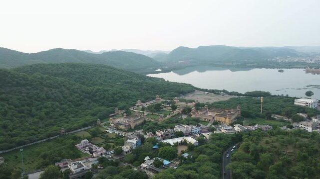 A wide aerial shot capturing the historic architecture of Jaipur, featuring Maota Lake and the lush green Aravalli mountain range during a hazy day.