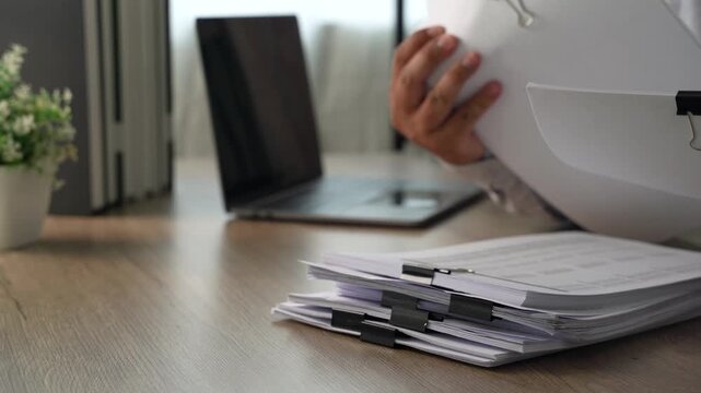 Person reviewing and turning stacked financial documents beside a laptop, showing accounting document review, report management and finance administration.