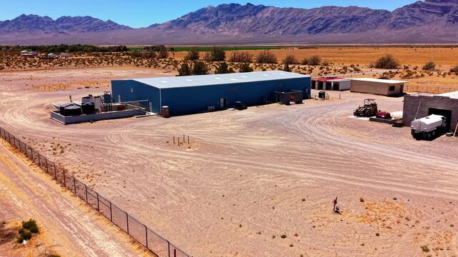 Stable aerial view surveys an isolated industrial building, equipment yard and surrounding desert terrain.