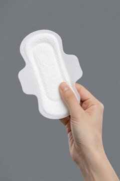 Close up of female hand holding white sanitary pad against gray background. Woman shows menstrual hygiene product for period care. Concept of feminine health, monthly cycle protection, personal