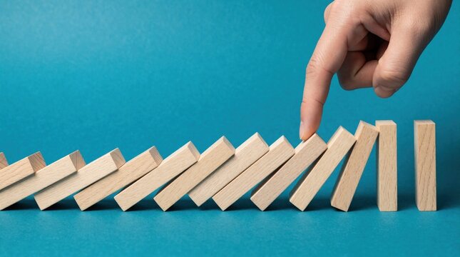 Human hand uses finger to stop falling wooden domino blocks on blue background. Person prevents domino effect, manages crisis. Business concept of risk control, stability, strategic decision making