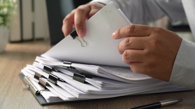 Hands sorting and turning stacked clipped documents on a desk, showing document management, file review and office administration workflow.