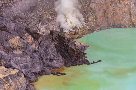 Ijen volcano complex in East Java, Indonesia, with a turquoise acidic crater lake, rugged rock walls, yellow sulfur deposits, and thick white smoke rising from active vents.
