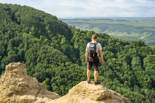 Man tourist with a backpack standing on a rocky, protruding ledge against scenic green hills and wooded slopes