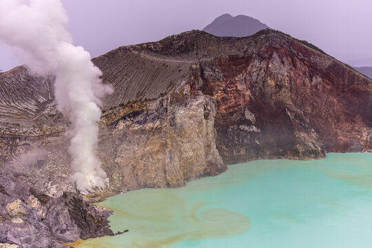 Ijen volcano complex in East Java, Indonesia, with a turquoise acidic crater lake, rugged rock walls, yellow sulfur deposits, and thick white smoke rising from active vents.