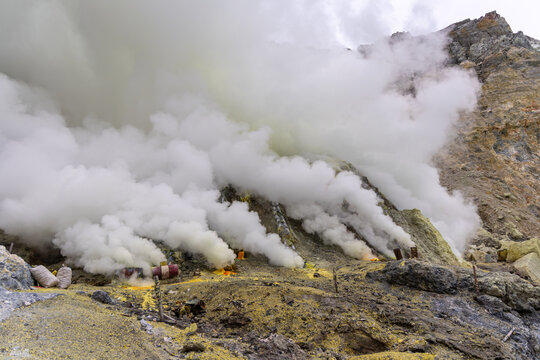 Ijen volcano complex in East Java, Indonesia, with a turquoise acidic crater lake, rugged rock walls, yellow sulfur deposits, and thick white smoke rising from active vents.