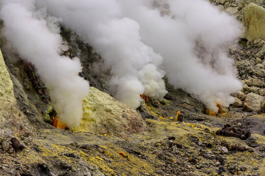 Ijen volcano complex in East Java, Indonesia, with a turquoise acidic crater lake, rugged rock walls, yellow sulfur deposits, and thick white smoke rising from active vents.