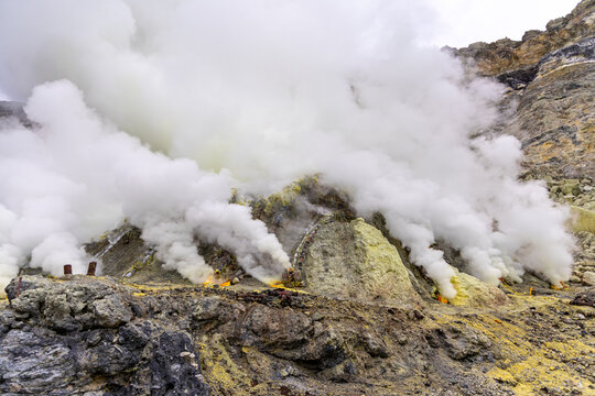 Ijen volcano complex in East Java, Indonesia, with a turquoise acidic crater lake, rugged rock walls, yellow sulfur deposits, and thick white smoke rising from active vents.