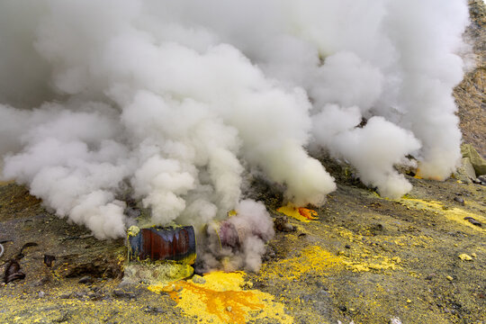 Ijen volcano complex in East Java, Indonesia, with a turquoise acidic crater lake, rugged rock walls, yellow sulfur deposits, and thick white smoke rising from active vents.