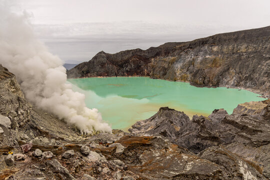 Ijen volcano complex in East Java, Indonesia, with a turquoise acidic crater lake, rugged rock walls, yellow sulfur deposits, and thick white smoke rising from active vents.