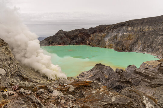 Ijen volcano complex in East Java, Indonesia, with a turquoise acidic crater lake, rugged rock walls, yellow sulfur deposits, and thick white smoke rising from active vents.