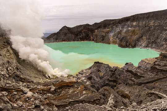 Ijen volcano complex in East Java, Indonesia, with a turquoise acidic crater lake, rugged rock walls, yellow sulfur deposits, and thick white smoke rising from active vents.