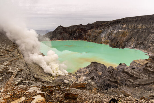 Ijen volcano complex in East Java, Indonesia, with a turquoise acidic crater lake, rugged rock walls, yellow sulfur deposits, and thick white smoke rising from active vents.
