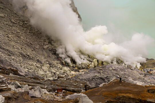 Ijen volcano complex in East Java, Indonesia, with a turquoise acidic crater lake, rugged rock walls, yellow sulfur deposits, and thick white smoke rising from active vents.
