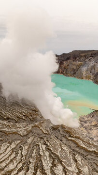 Ijen volcano complex in East Java, Indonesia, with a turquoise acidic crater lake, rugged rock walls, yellow sulfur deposits, and thick white smoke rising from active vents.